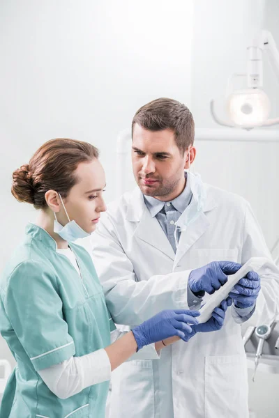 Dentiste masculin tenant tablette numérique et regardant collègue attrayant dans la clinique dentaire — Photo de stock