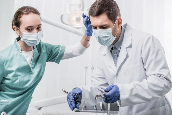 Dentista feminina em máscara de pé perto colega segurando instrumentos dentários — Fotografia de Stock