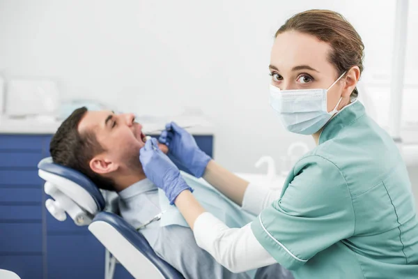 Foco seletivo do dentista olhando para a câmera e segurando instrumentos dentários perto do paciente com boca aberta — Fotografia de Stock