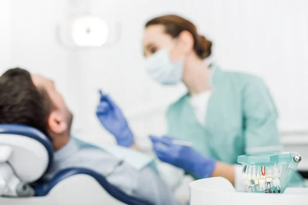 Messa a fuoco selettiva del modello dentale vicino al paziente e al dentista in maschera con strumenti dentali — Stock Photo