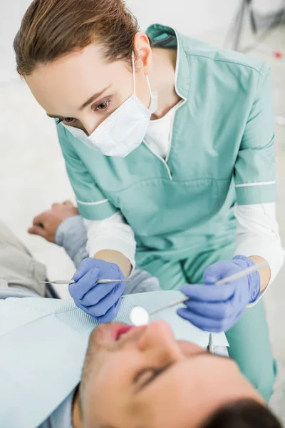 Enfoque selectivo del dentista femenino en la máscara que sostiene los instrumentos dentales y examinar al paciente - foto de stock