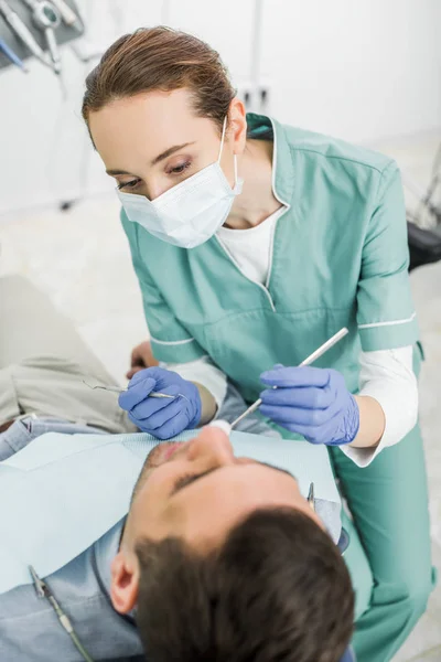 Dentista femenina con máscara que sostiene instrumentos dentales y examina a la paciente en la clínica dental - foto de stock