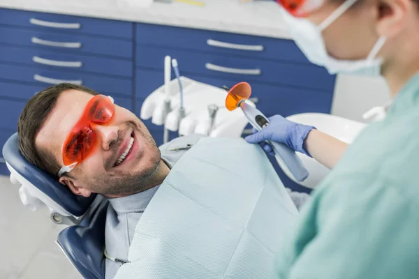 Selective focus of cheerful man in glasses near dentist holding dental tool before bleaching procedure — Stock Photo