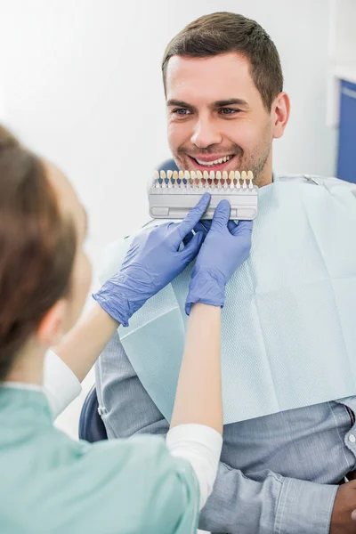 Messa a fuoco selettiva di uomo allegro sorridente vicino dentista con tavolozza di colori di denti in mani — Foto stock