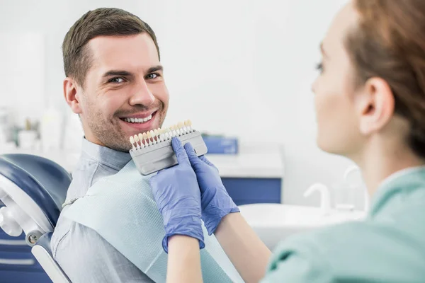 Selektiver Fokus des glücklichen Mannes, der in der Nähe des Zahnarztes lächelt, mit der Farbpalette der Zähne in den Händen — Stockfoto