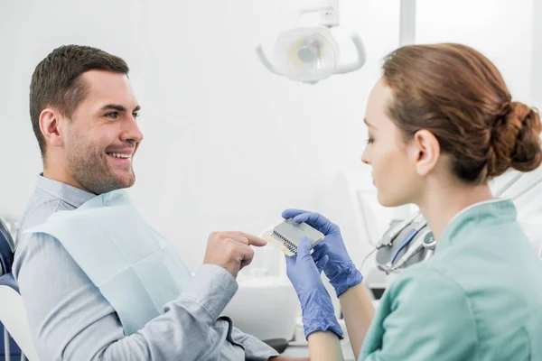 Щасливий чоловік вказує пальцем на кольорову палітру зубів в руках стоматолога — стокове фото