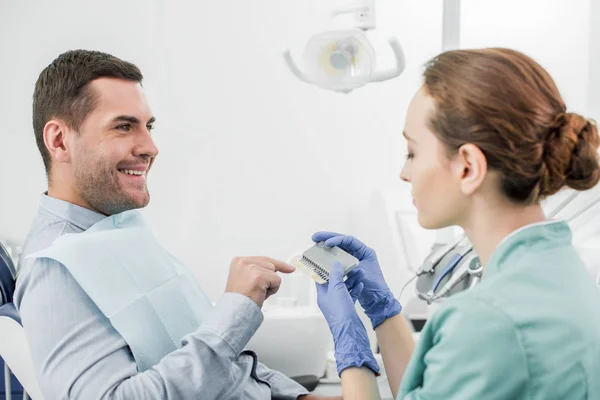 Homem feliz apontando com o dedo na paleta de cores dos dentes nas mãos do dentista — Fotografia de Stock