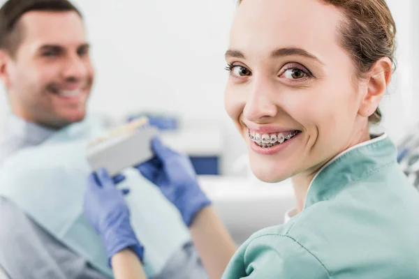 Enfoque selectivo de dentista alegre con frenos en los dientes sonriendo mientras sostiene la paleta de colores cerca del paciente - foto de stock