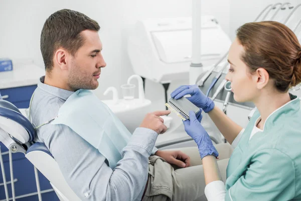 Beau patient pointant du doigt la palette de couleurs des dents près du dentiste — Photo de stock