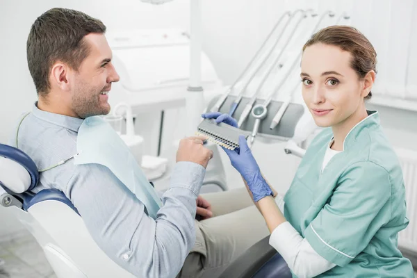 Paciente feliz apuntando con el dedo a la paleta de colores de los dientes cerca del dentista - foto de stock