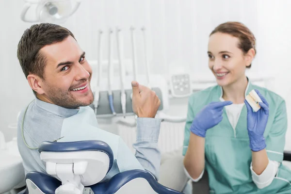 Selective focus of handsome man showing thumb up near beautiful dentist holding teeth model — Stock Photo