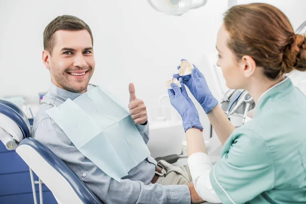 Enfoque selectivo del hombre feliz mostrando el pulgar hacia arriba cerca del dentista femenino sosteniendo el modelo de dientes - foto de stock