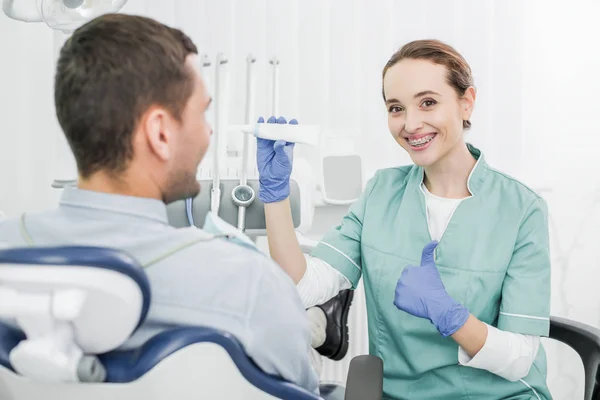 Selektiver Fokus der fröhlichen Zahnärztin, die Zahnpasta in der Hand hält und den Daumen in die Nähe der Patientin zeigt — Stockfoto