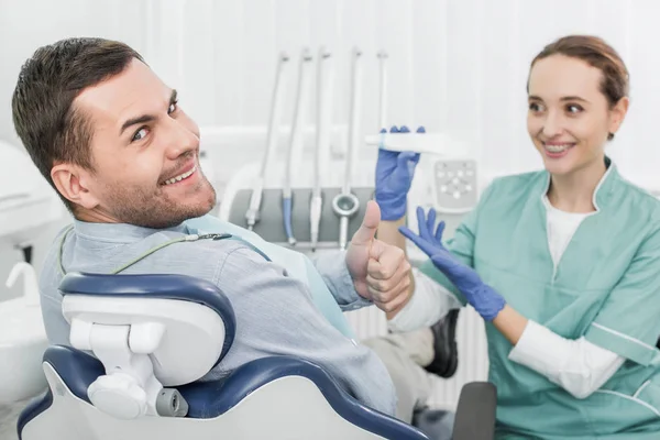 Enfoque selectivo de paciente alegre mostrando el pulgar hacia arriba cerca del dentista femenino sosteniendo pasta de dientes - foto de stock
