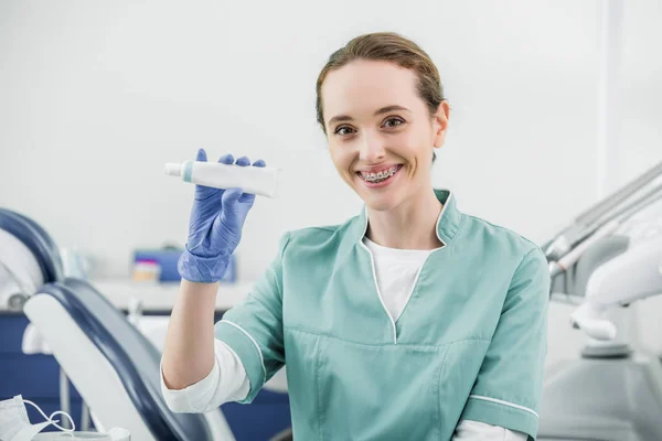 Cheerful female dentist with braces on teeth holding toothpaste — Stock Photo