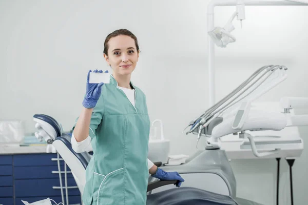 Hermoso dentista sosteniendo la tarjeta en blanco mientras está de pie en la clínica dental - foto de stock