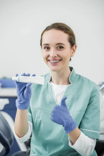 Dentiste féminine gaie avec des bretelles sur les dents tenant dentifrice et montrant pouce vers le haut — Photo de stock