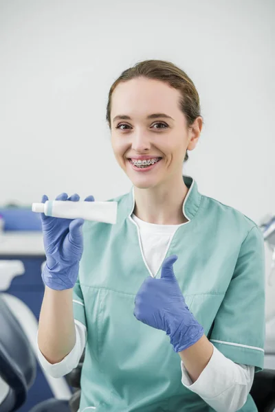 Dentista femenina alegre con frenos en los dientes sosteniendo la pasta de dientes y mostrando el pulgar hacia arriba - foto de stock