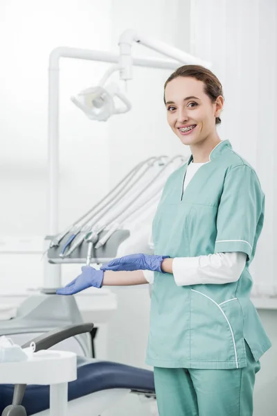 Привлекательный стоматолог с брекетами на зубах, улыбающийся во время жеста в стоматологической клинике — стоковое фото