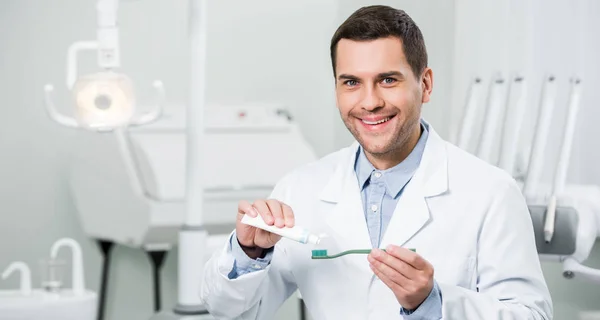 Dentista feliz espremendo pasta de dentes na escova de dentes na clínica odontológica — Fotografia de Stock