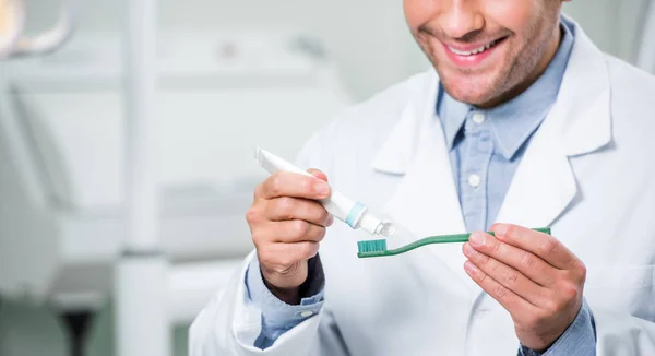 Vue recadrée du dentiste masculin heureux serrant le dentifrice sur la brosse à dents dans la clinique dentaire — Photo de stock