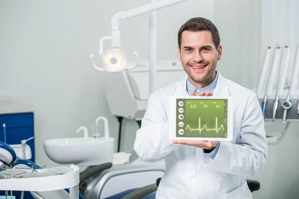 Alegre dentista sosteniendo tableta digital con aplicación médica en la pantalla en la clínica dental - foto de stock