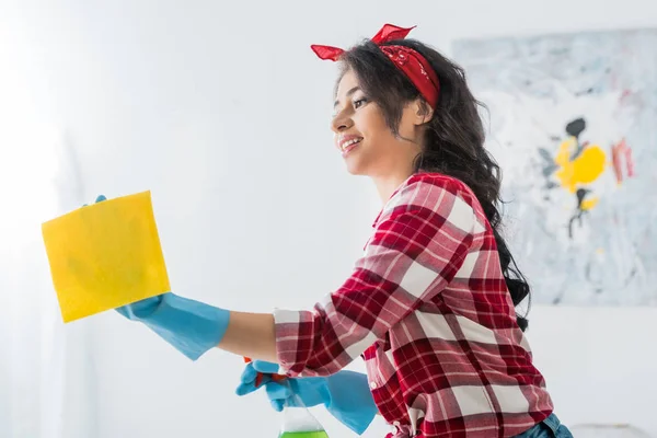 Attractive african american woman in plaid shirt and blue rubber gloves cleaning house with yellow rag — Stock Photo