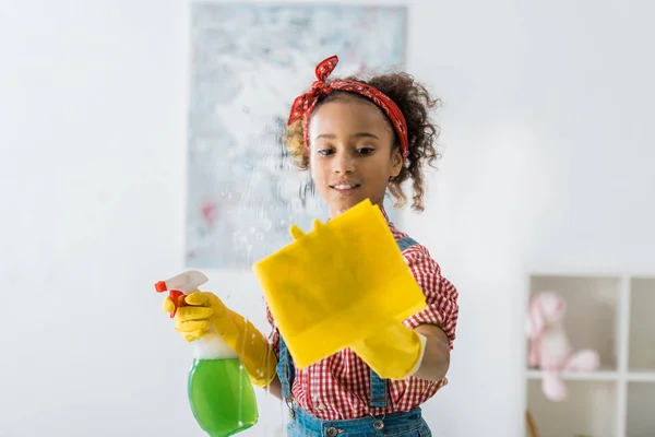 Mignon enfant afro-américain tenant chiffon jaune et bouteille de pulvérisation verte — Photo de stock