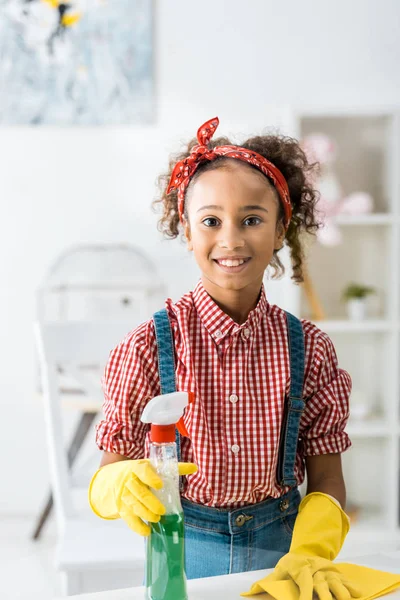 Smiling african american child with green spray bottle and yellow rag looking at camera — Stock Photo