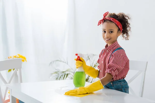 Cute african american child in plaid shirt cleaning table in yellow rubber gloves — Stock Photo