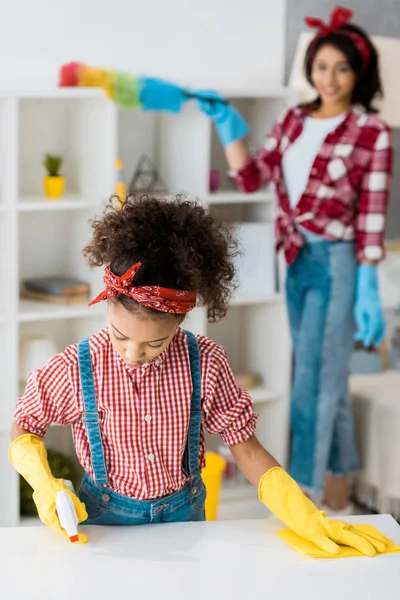 Foco seletivo de mesa de limpeza infantil afro-americana bonito enquanto a mãe polvilhar unidade de prateleira — Fotografia de Stock
