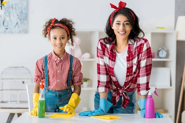 Feliz africana americana madre con adorable hija en brillante goma guantes y aerosol botellas limpieza casa - foto de stock