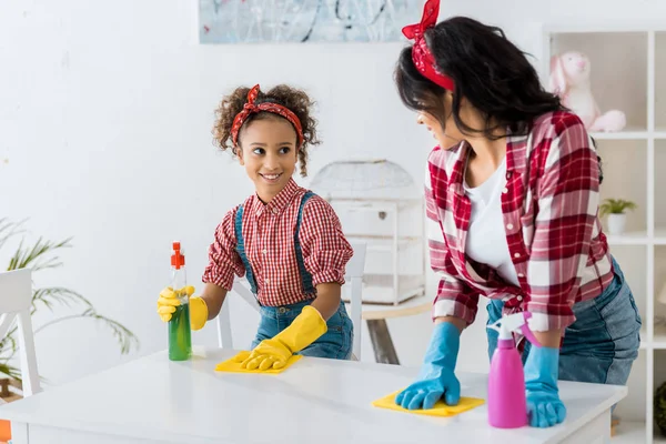 Souriant afro-américain maman et fille nettoyage table blanche avec chiffons jaunes — Photo de stock