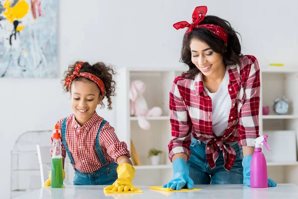 Feliz afro-americana mãe com adorável filha mesa de limpeza em luvas de borracha brilhantes — Fotografia de Stock