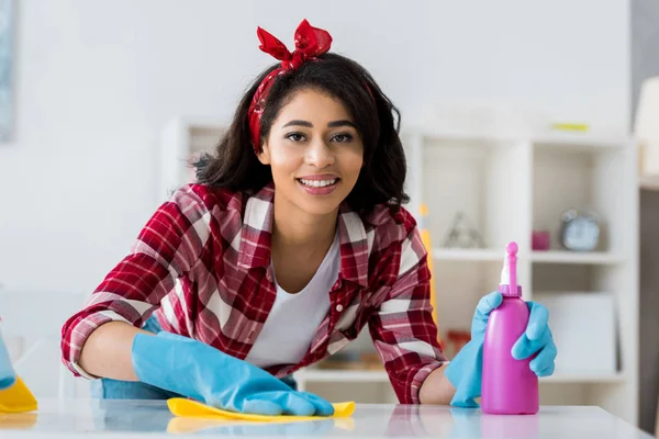 Sorrindo mulher americana africana com pano amarelo e frasco de spray rosa olhando para a câmera — Fotografia de Stock