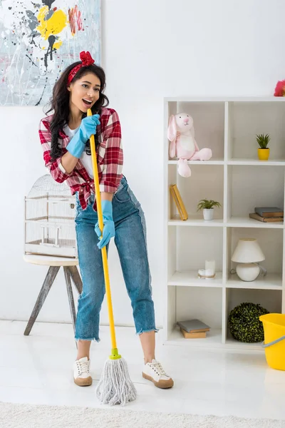 Beautiful african american woman in plaid shirt and blue rubber gloves singing with mop — Stock Photo