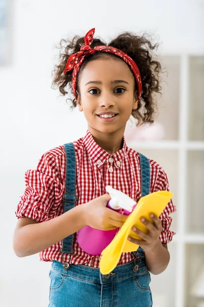 Adorable african american child spraying cleaning liquid on yellow rag and looking at camera — Stock Photo