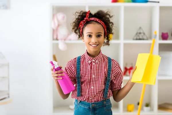 Sonriente afroamericana chica sosteniendo botella de spray rosa y trapo amarillo mientras mira la cámara - foto de stock