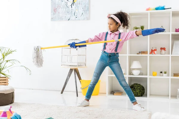Cute african american girl in blue rubber gloves dancing with mop — Stock Photo