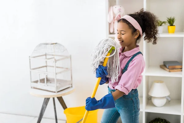 Cute african american girl in blue rubber gloves singing with broom — Stock Photo