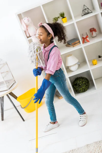 Cute african american child in pink shirt and blue jeans singing with mop — Stock Photo