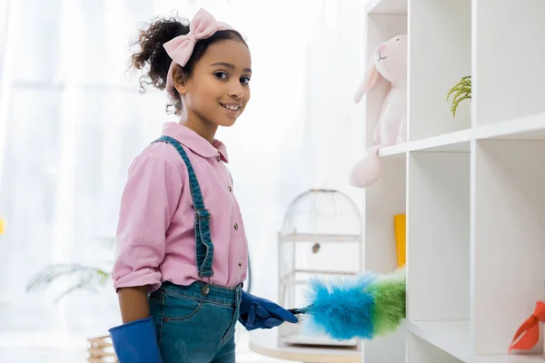 Adorable african american child cleaning shelves with duster — Stock Photo