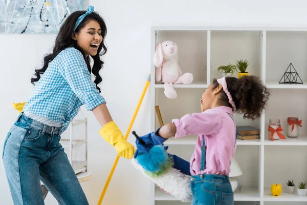 Laughing african american mother with cute daughter having fun while fighting with dusters — Stock Photo