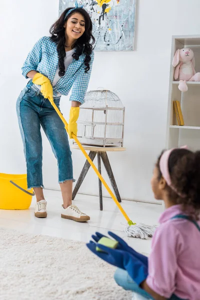 Smiling african american woman washing floor with mop while daughter sitting on carpet — Stock Photo