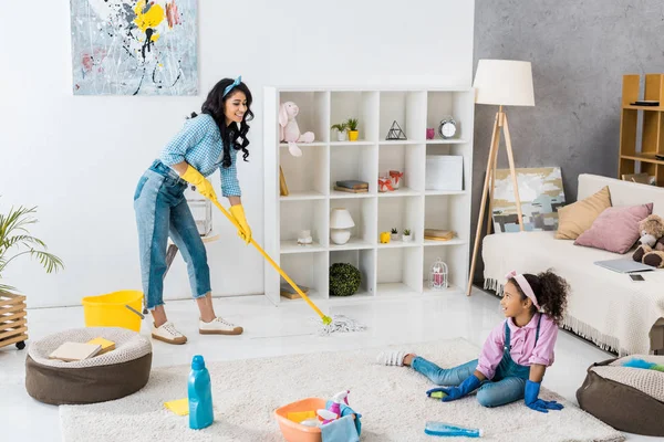 Гарна афроамериканка миє підлогу, дочка прибирає килим — стокове фото