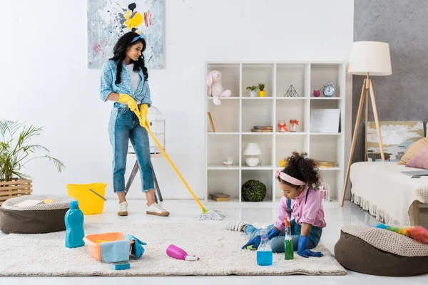 Lindo africano americano niño limpieza alfombra mientras madre lavado piso con fregona - foto de stock