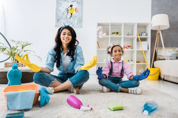 Sorridente afro-americano mãe e filha em luvas de borracha brilhante sentado no tapete em poses de lótus — Fotografia de Stock