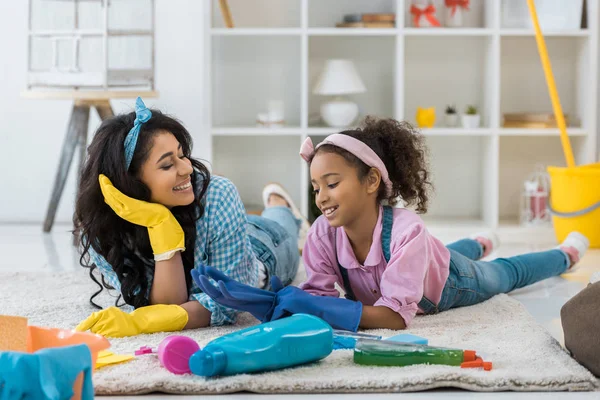 Smiling african american mom and cute child in bright rubber gloves lying on carpet — Stock Photo
