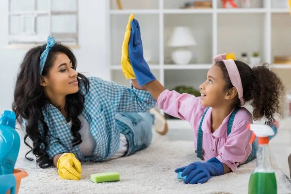 African american mom and daughter in bright rubber gloves giving high five while lying on carpet — Stock Photo