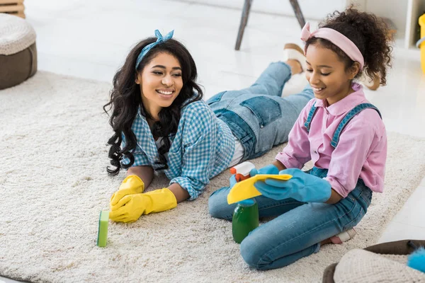 Sonriente afroamericana mujer acostada en la alfombra mientras hija sentada cerca - foto de stock